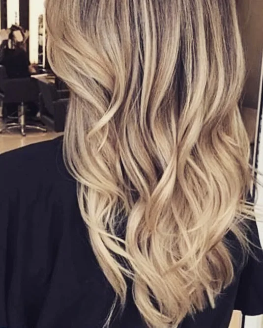 Blonde balayage waves down the back, blurred salon environment behind — colour by Jolin